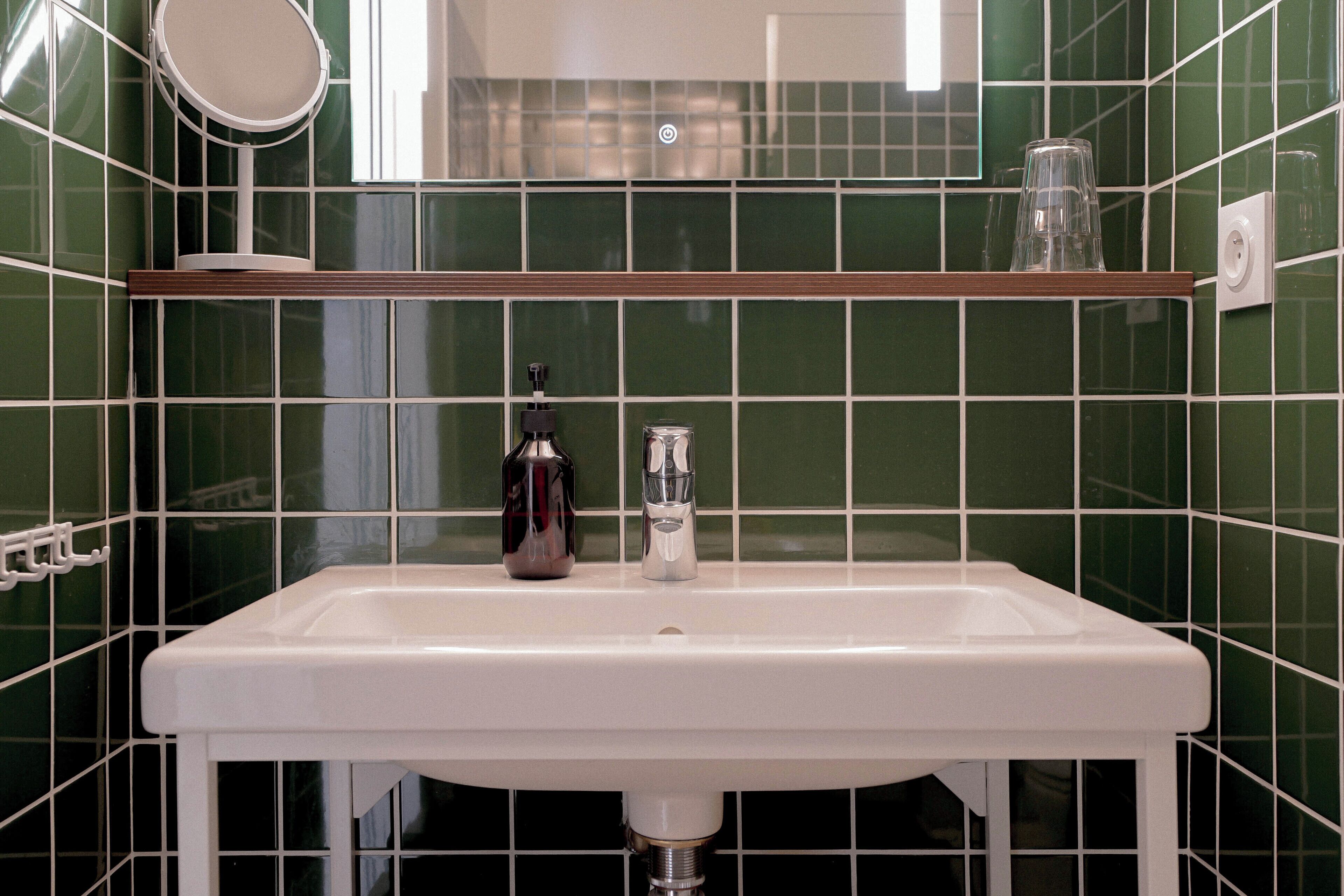 Un baño con un lavabo blanco, paredes con azulejos verdes, un espejo y una repisa con un vaso y una botella.