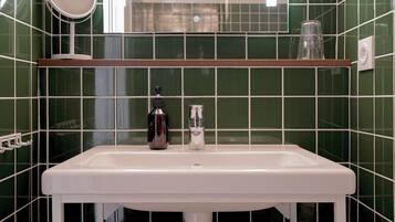 Un baño con un lavabo blanco, paredes con azulejos verdes, un espejo y una repisa con un vaso y una botella.
