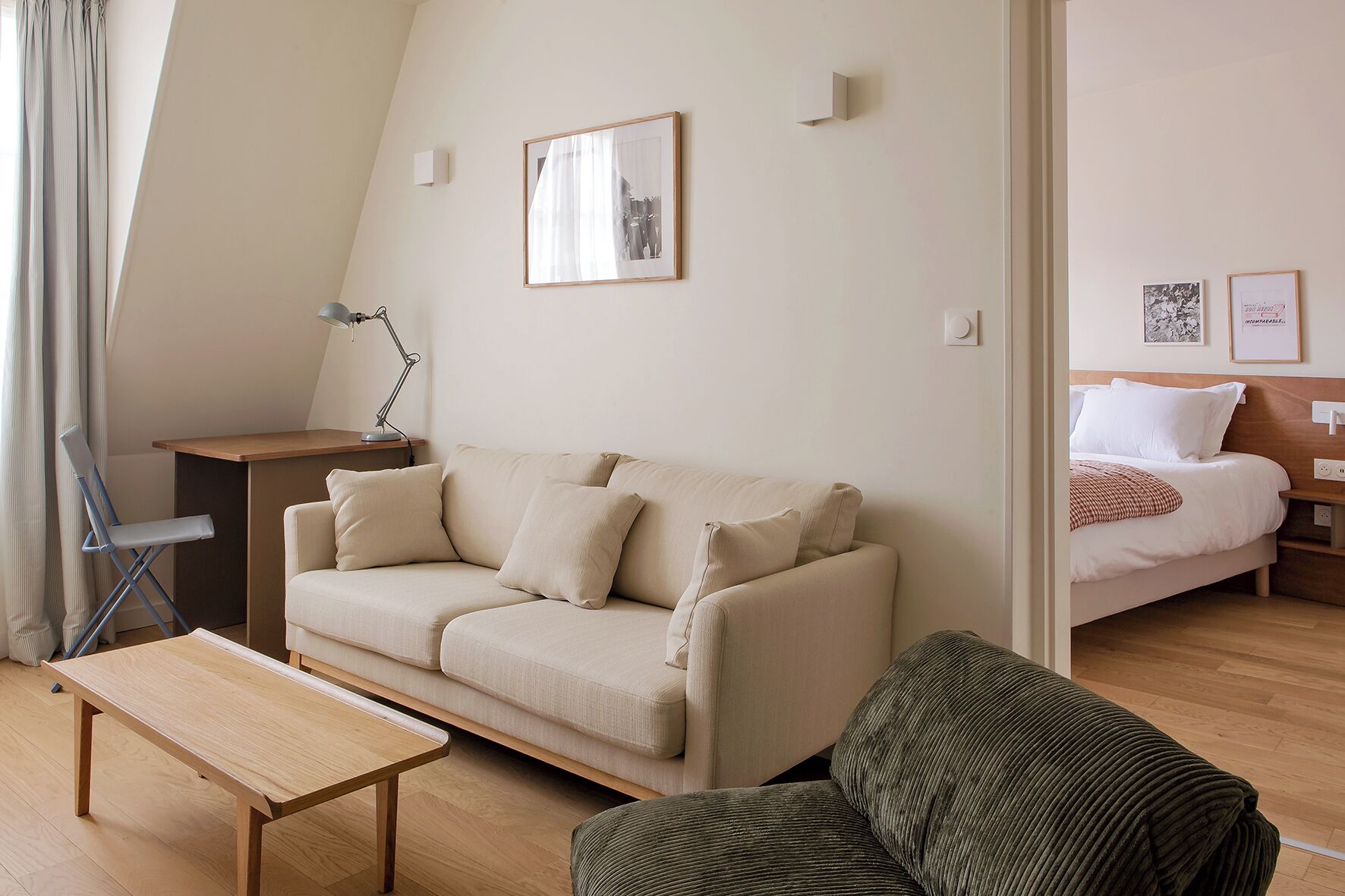 Habitación moderna y bien iluminada con un sofá beige, una mesita de madera pequeña y una silla verde. Se ve una cama con ropa blanca de cama al fondo.