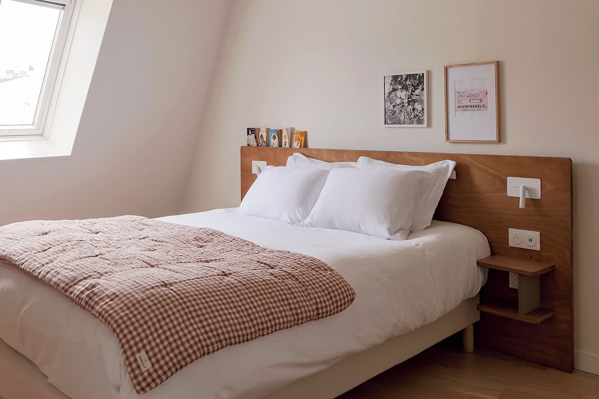 Una cama bien hecha con una colcha a cuadros, almohadas blancas y un cabecero de madera. Hay dos cuadros enmarcados en la pared, una mesita de noche con una lámpara y una ventana con vistas a edificios.