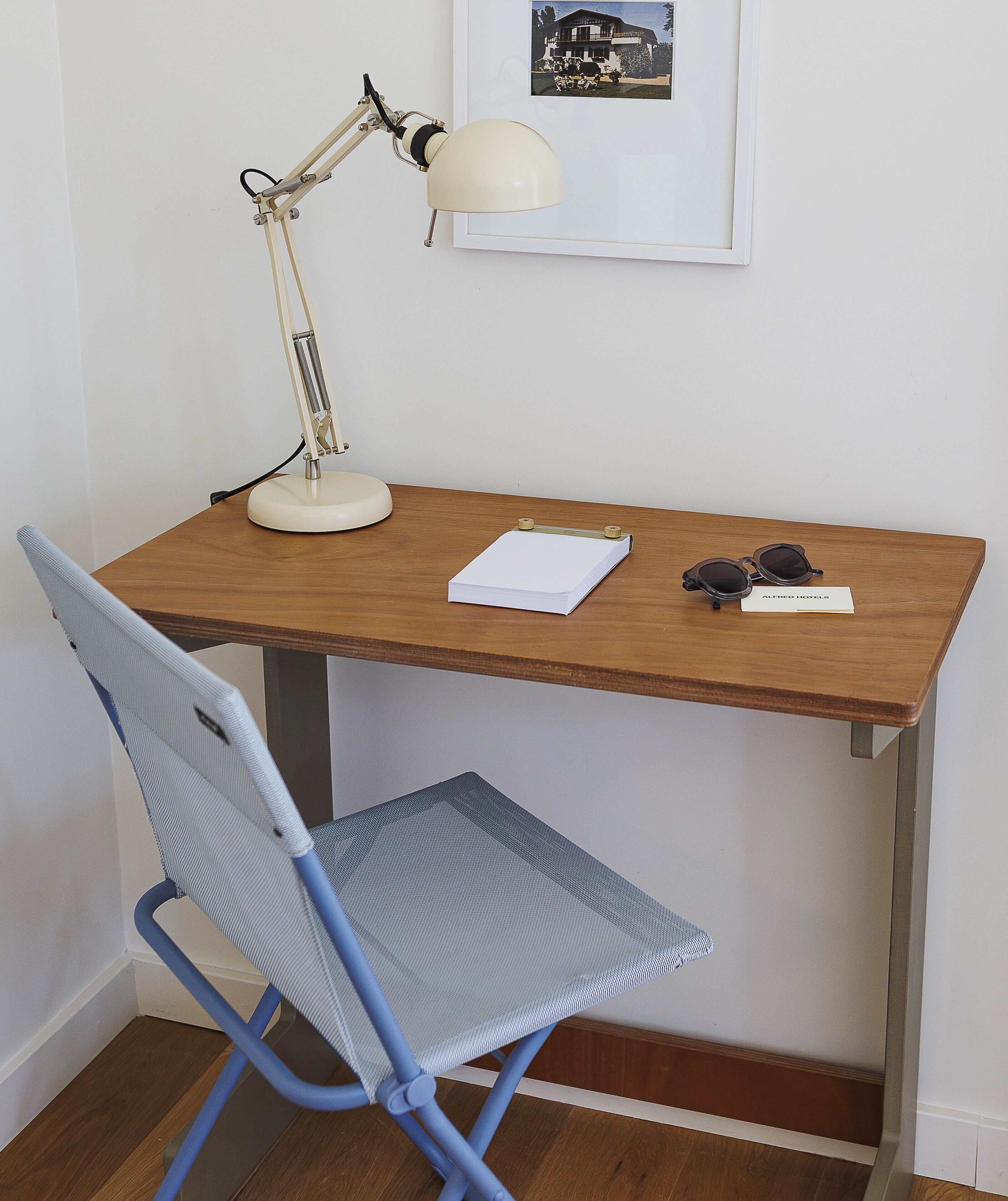 Un bureau en bois, une lampe de bureau blanche, une chaise bleue, un bloc-notes et une paire de lunettes de soleil.