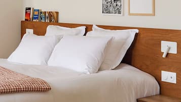 Una cama bien hecha con ropa de cama blanca, una mesita de noche pequeña y un enchufe en la pared.