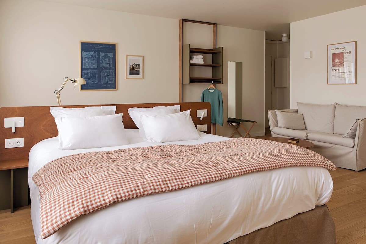 Habitación de hotel con una cama grande, una mesita de noche, una lámpara, un sofá, una pequeña mesa y una pared con cuadros.