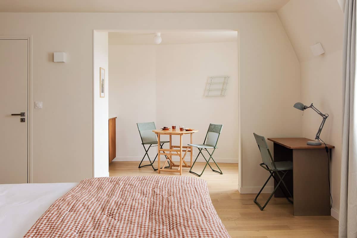 Un dormitorio con cama, escritorio y silla. En el fondo, una mesa con sillas.