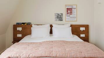 Een net opgemaakt bed met een geruit plaid, twee witte kussens en een houten hoofdbord. Twee inbouwschakelaars en een ingelijste poster sieren de muur.