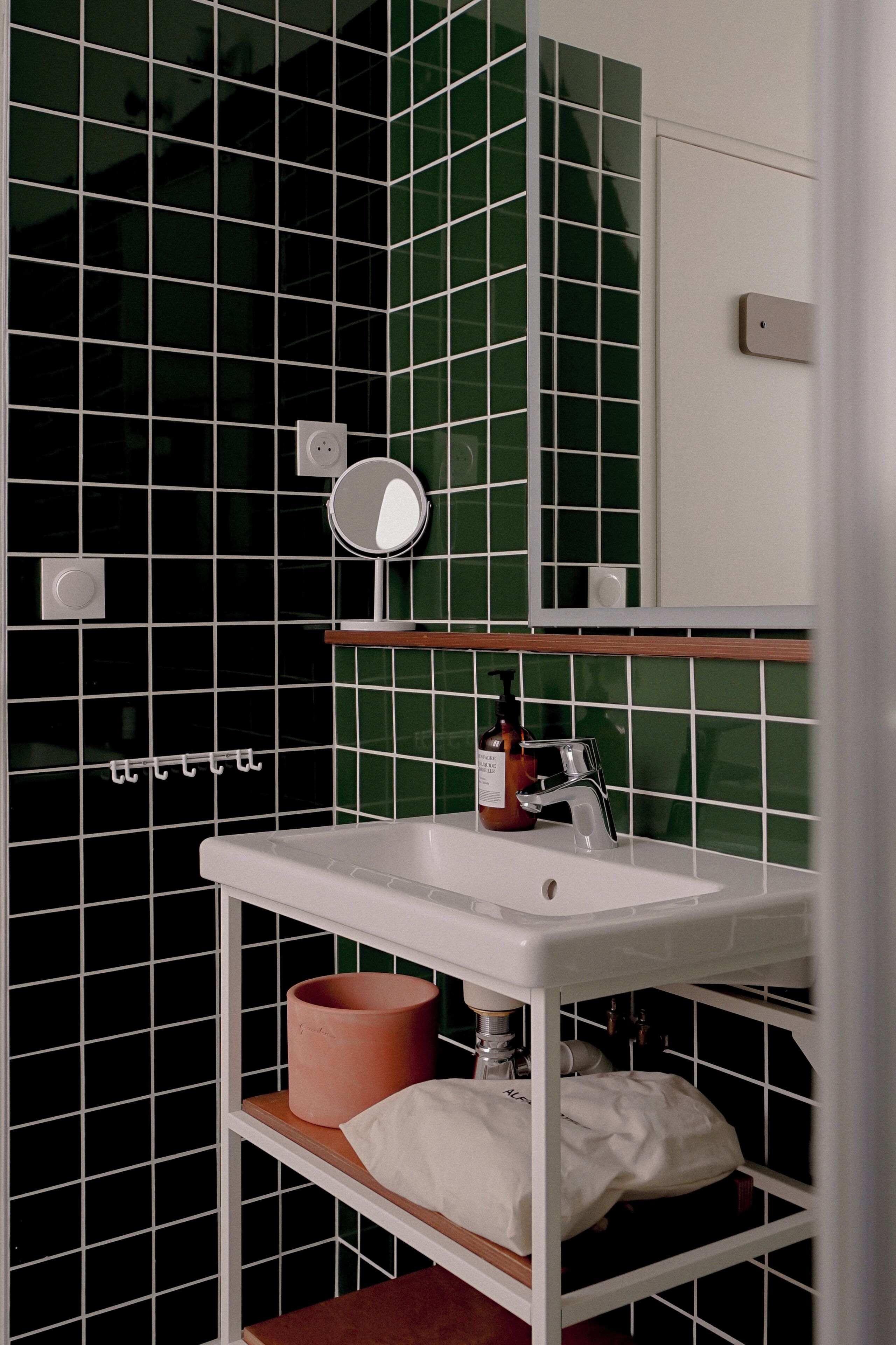 Een badkamer met een witte wastafel, groene betegelde wanden, een spiegel en een plank met een handdoek en een zeepdispenser.