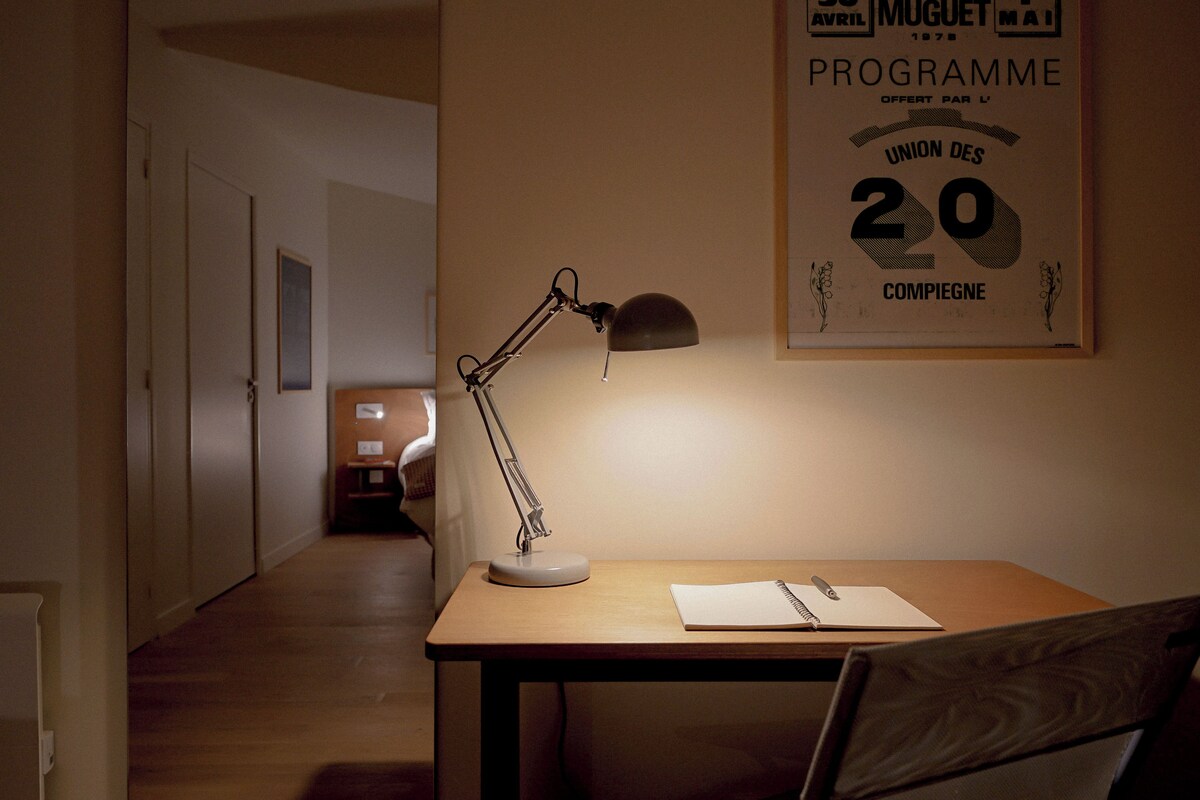 A desk with a lamp, a notepad, and a pen. There is a framed poster on the wall.