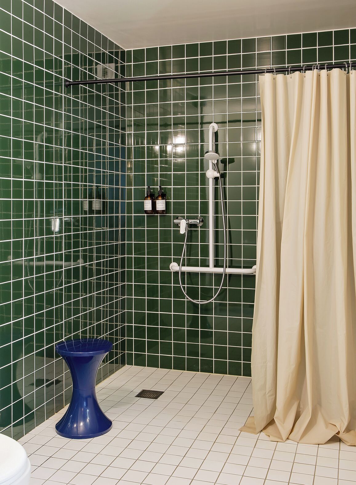 Une salle de bain avec des murs recouverts de carreaux verts, un espace douche, un rideau beige et un lavabo sur pied bleu.