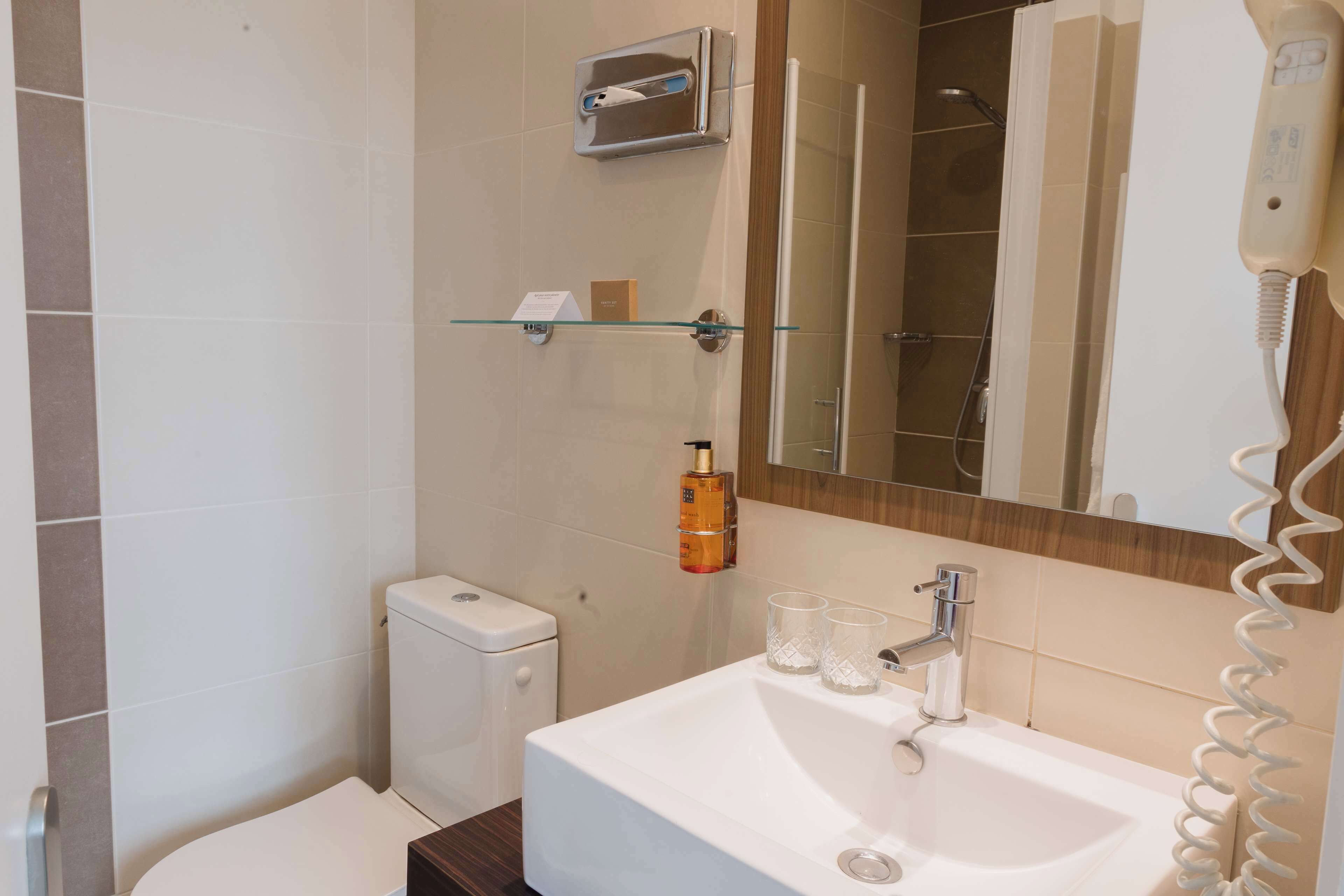 Un baño con lavamanos blanco, dispensador de jabón de vidrio y secador de pelo de mano.