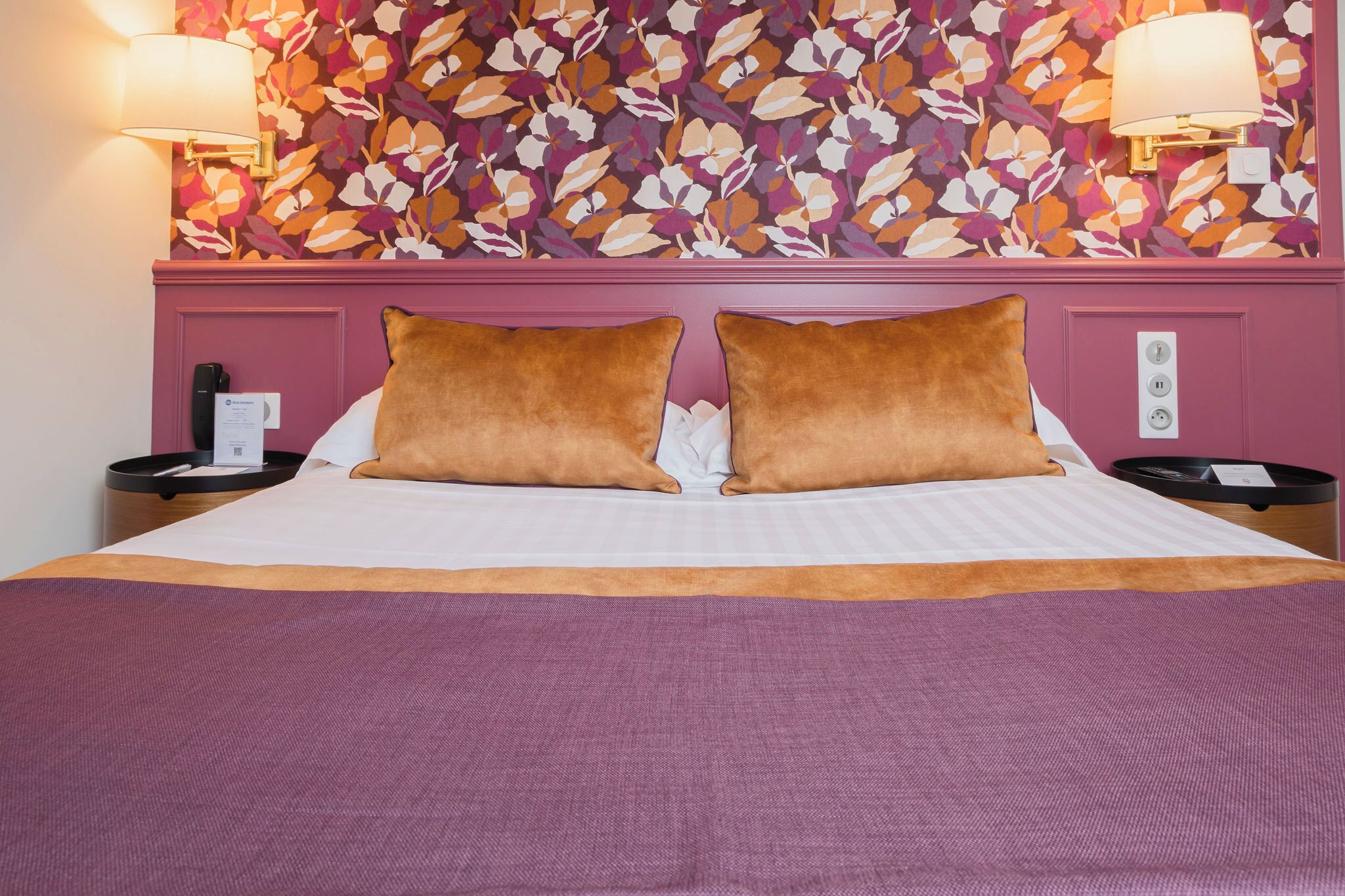 Una habitación de hotel con una cama, dos almohadas, una mesita de noche, una lámpara y un papel tapiz con motivos florales.