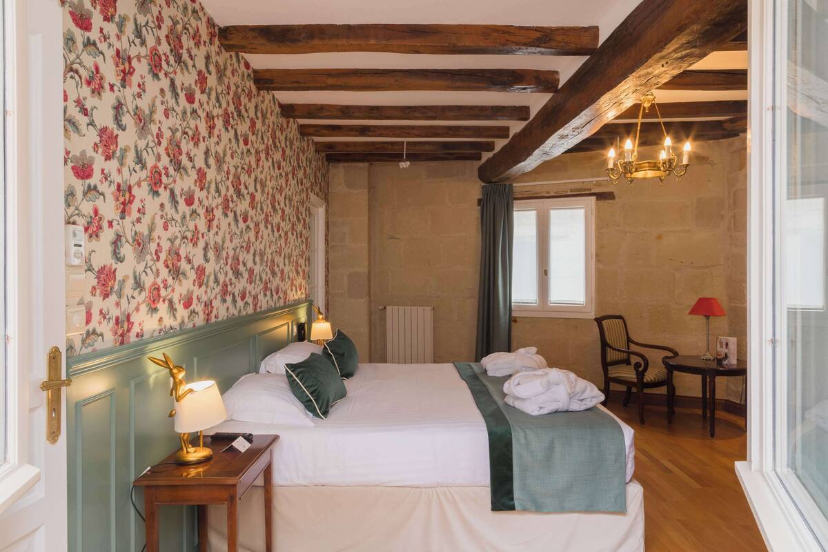 Une chambre d’hôtel avec deux lits, un papier peint à motifs floraux, des poutres en bois, un lustre, une table avec une lampe rouge et une chaise.