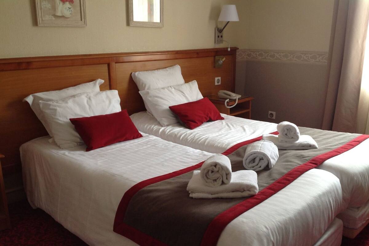 Une chambre d’hôtel avec deux lits, du linge de lit blanc, des oreillers rouges, une tête de lit en bois, une table de chevet avec une lampe et des serviettes roulées posées sur les lits.