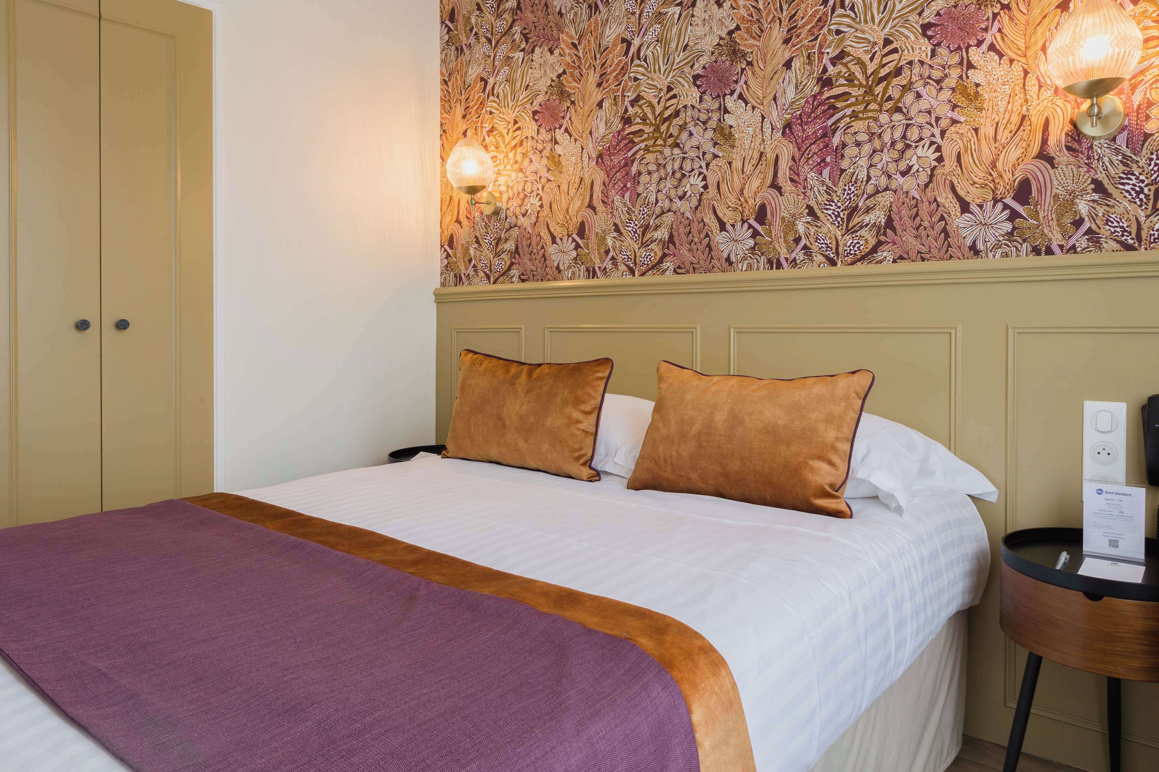 Habitación de hotel con cama, dos almohadas, mesita de noche con lámpara y pared con estampado floral.