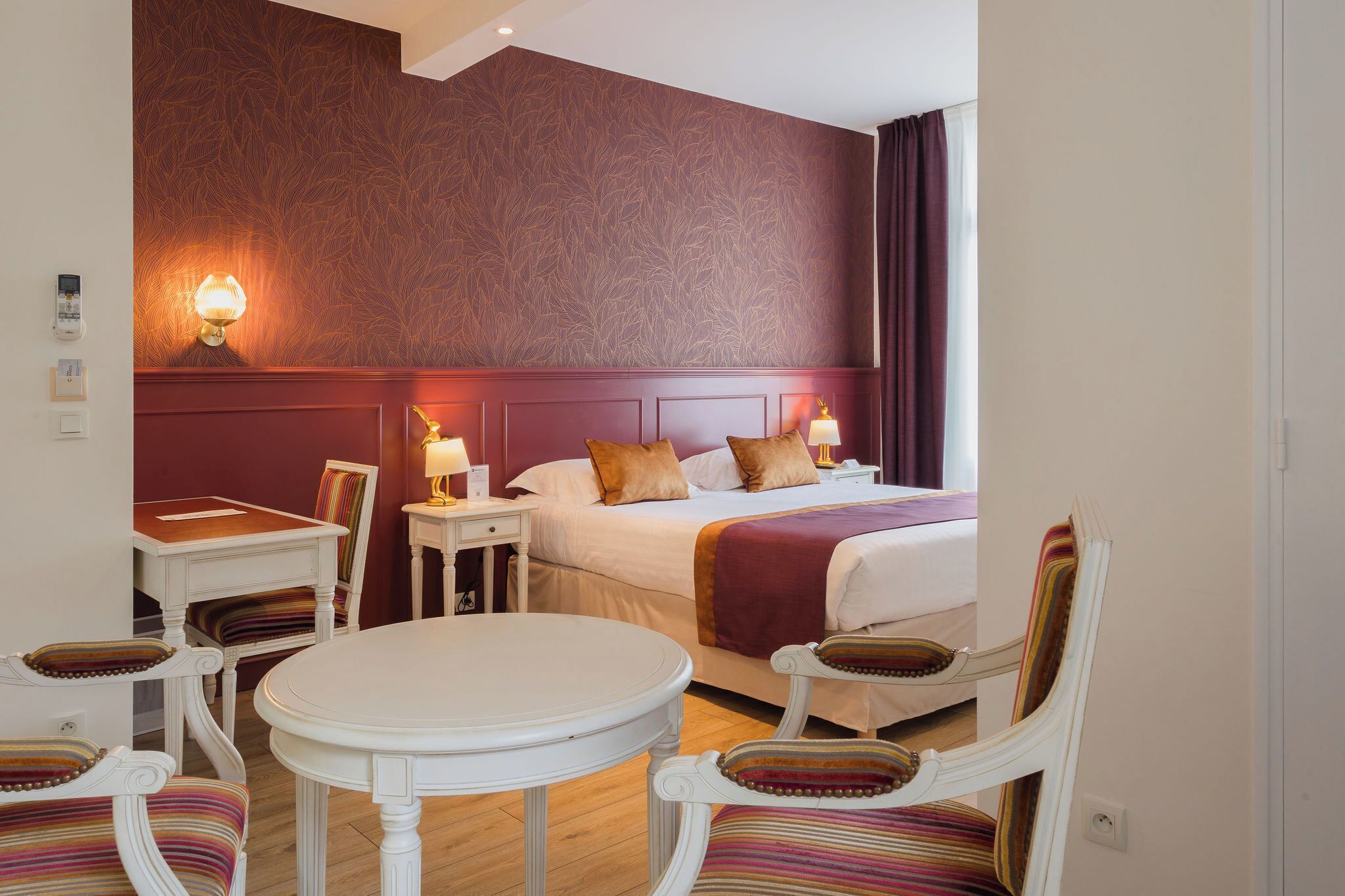 A hotel room with a bed, two bedside tables, a round table, and striped chairs.