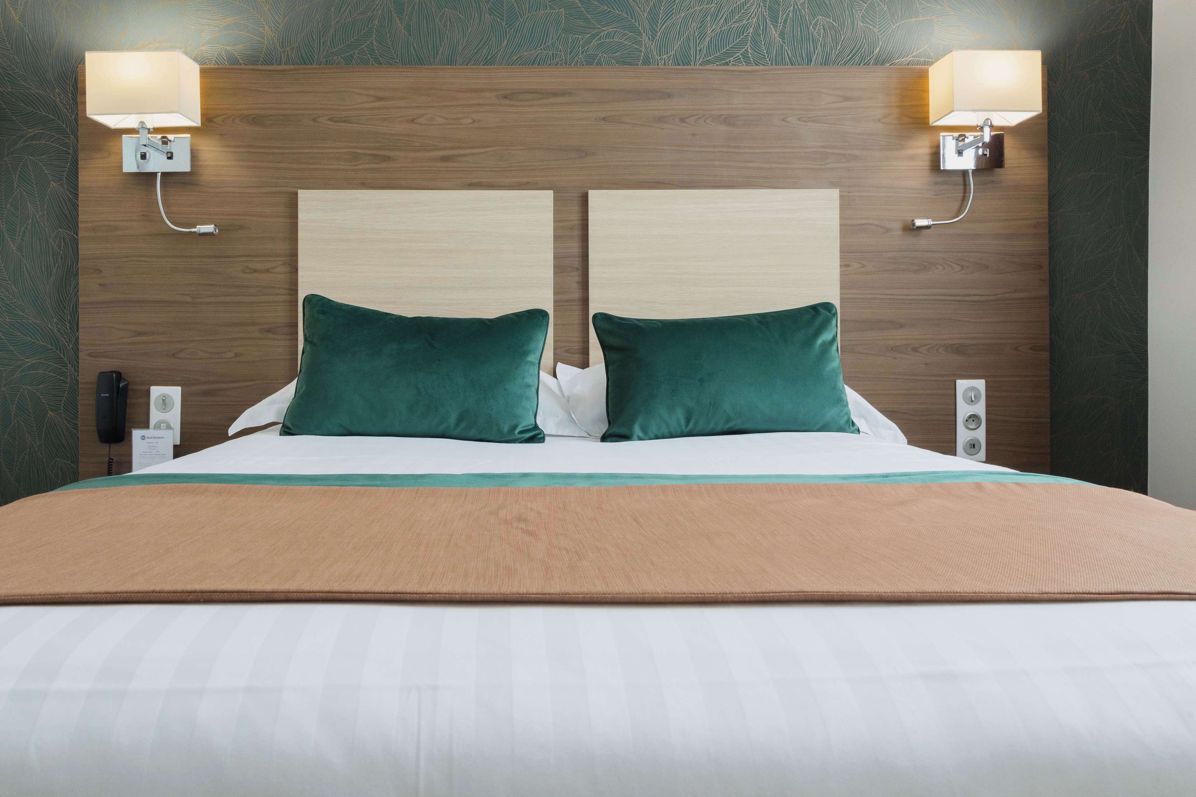 A hotel room with a wooden headboard, two green pillows, a white bedspread, and two wall-mounted lamps.
