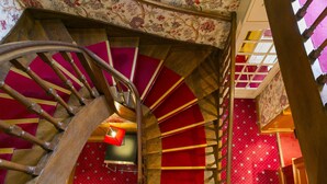 A spiral staircase with wooden steps and a metal handrail, leading to a red-carpeted area with patterned wallpaper.