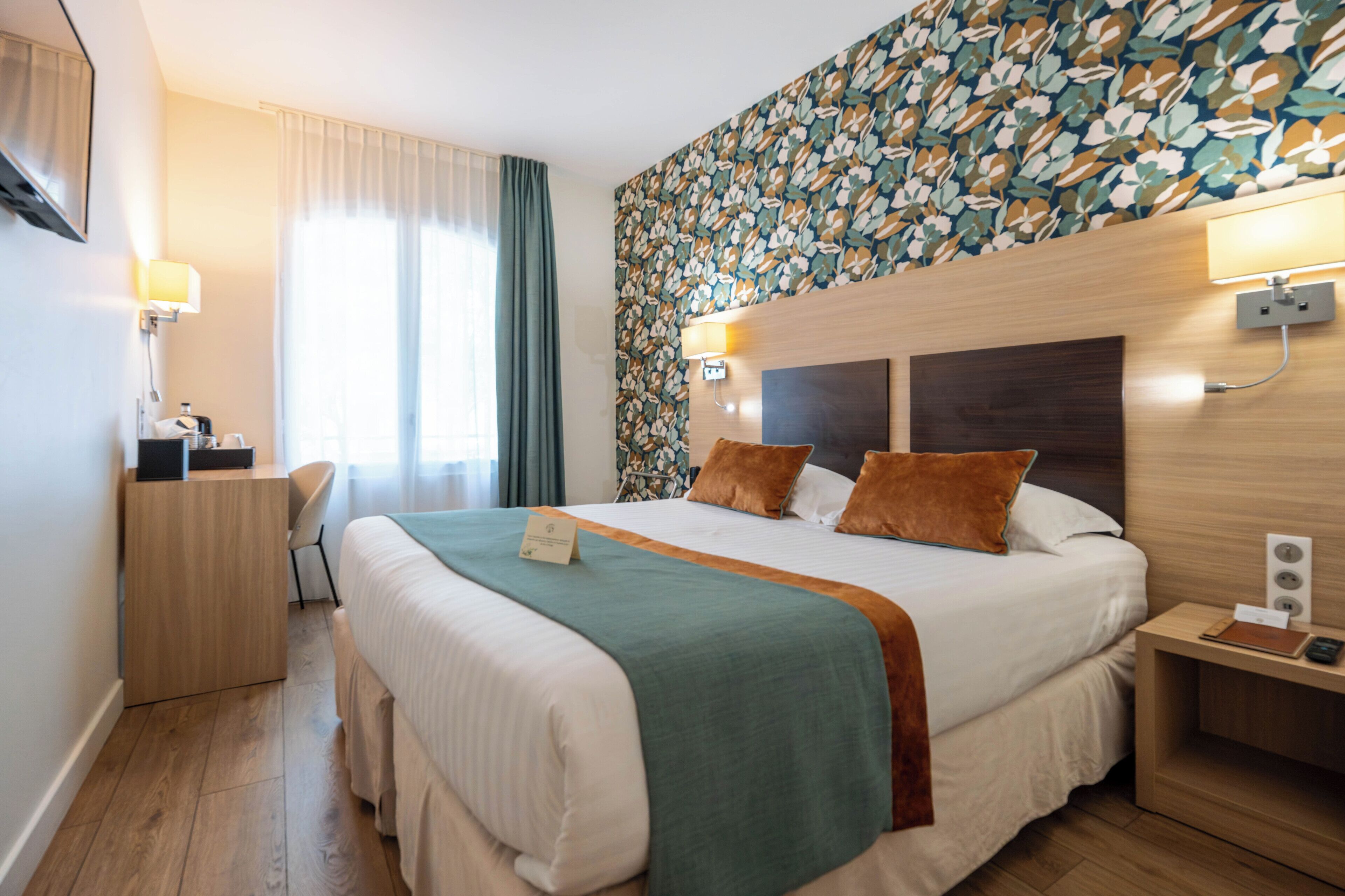 A hotel room with a large bed, a desk, a chair, a nightstand, and a wall with a floral pattern.