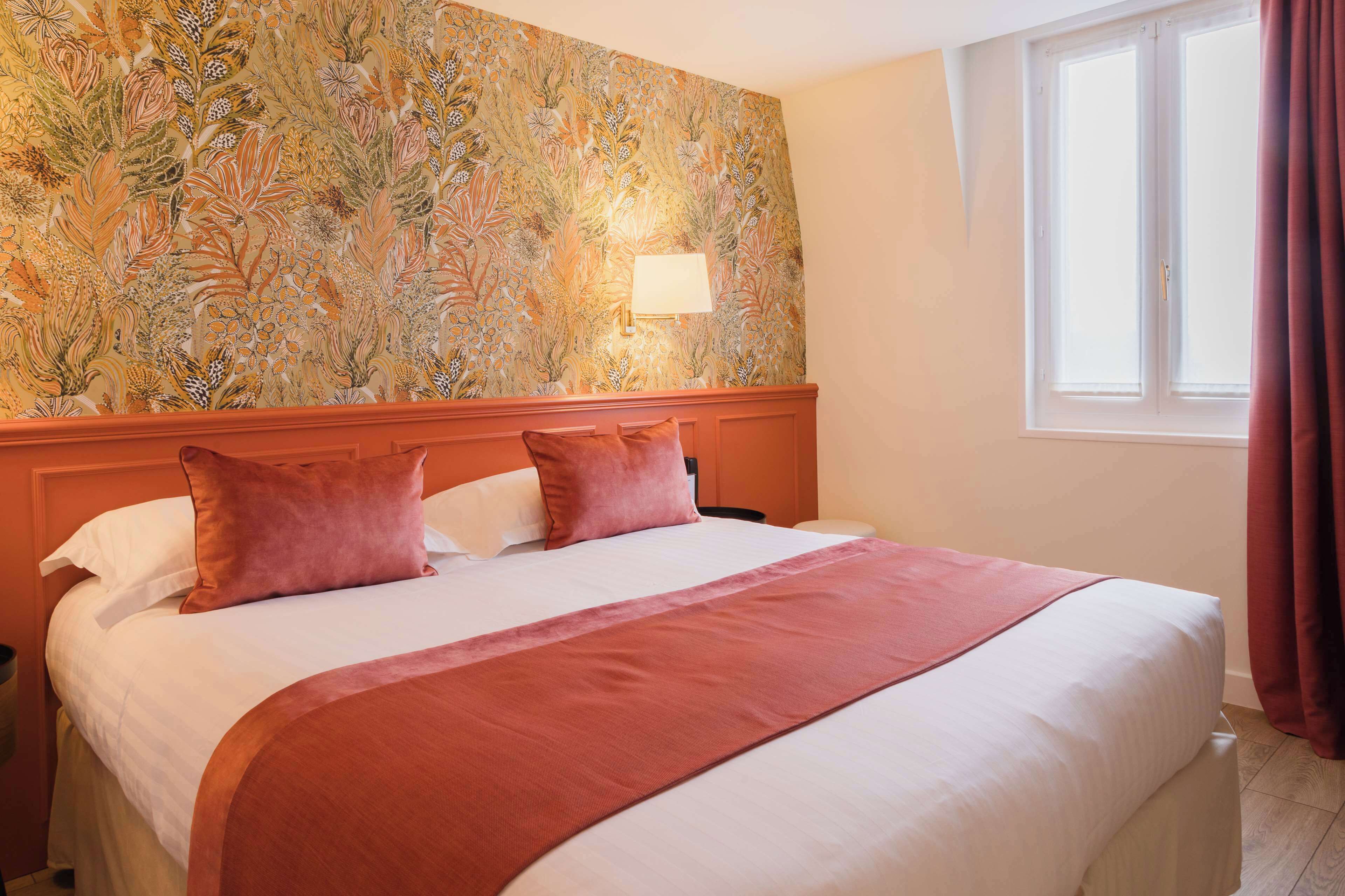 Habitación de hotel con una cama grande, papel tapiz floral, una mesita de noche y un ventanal con cortinas.