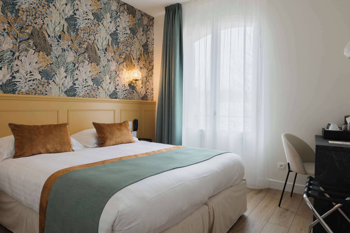 Une chambre d’hôtel avec un grand lit, un bureau et une chaise. Il y a une fenêtre avec des rideaux et un mur orné d’un motif floral.