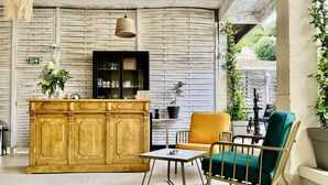 A wooden bar area with a green cushioned chair and a small table, a hanging light fixture, and a potted plant.