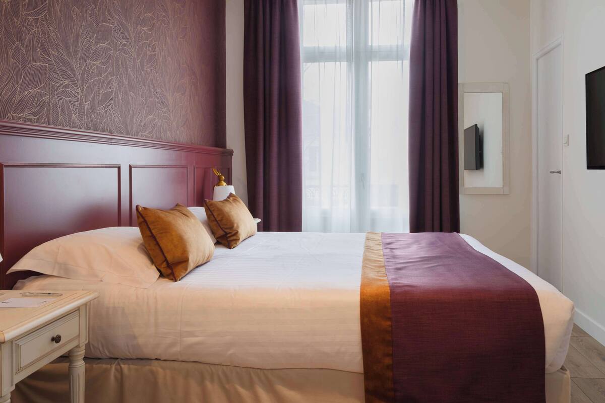 Un lit bien fait, avec du linge de lit blanc et un plaid bordeaux, une lampe de chevet et une table de chevet avec un téléphone.