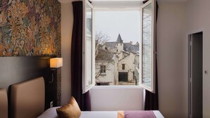 A hotel room with a bed, a nightstand with a lamp, a window with a view of buildings, and a wall with a floral pattern.