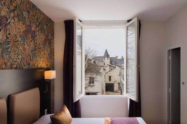 Habitación de hotel con cama, mesita de noche con lámpara, ventana con vista a edificios y pared con estampado floral.