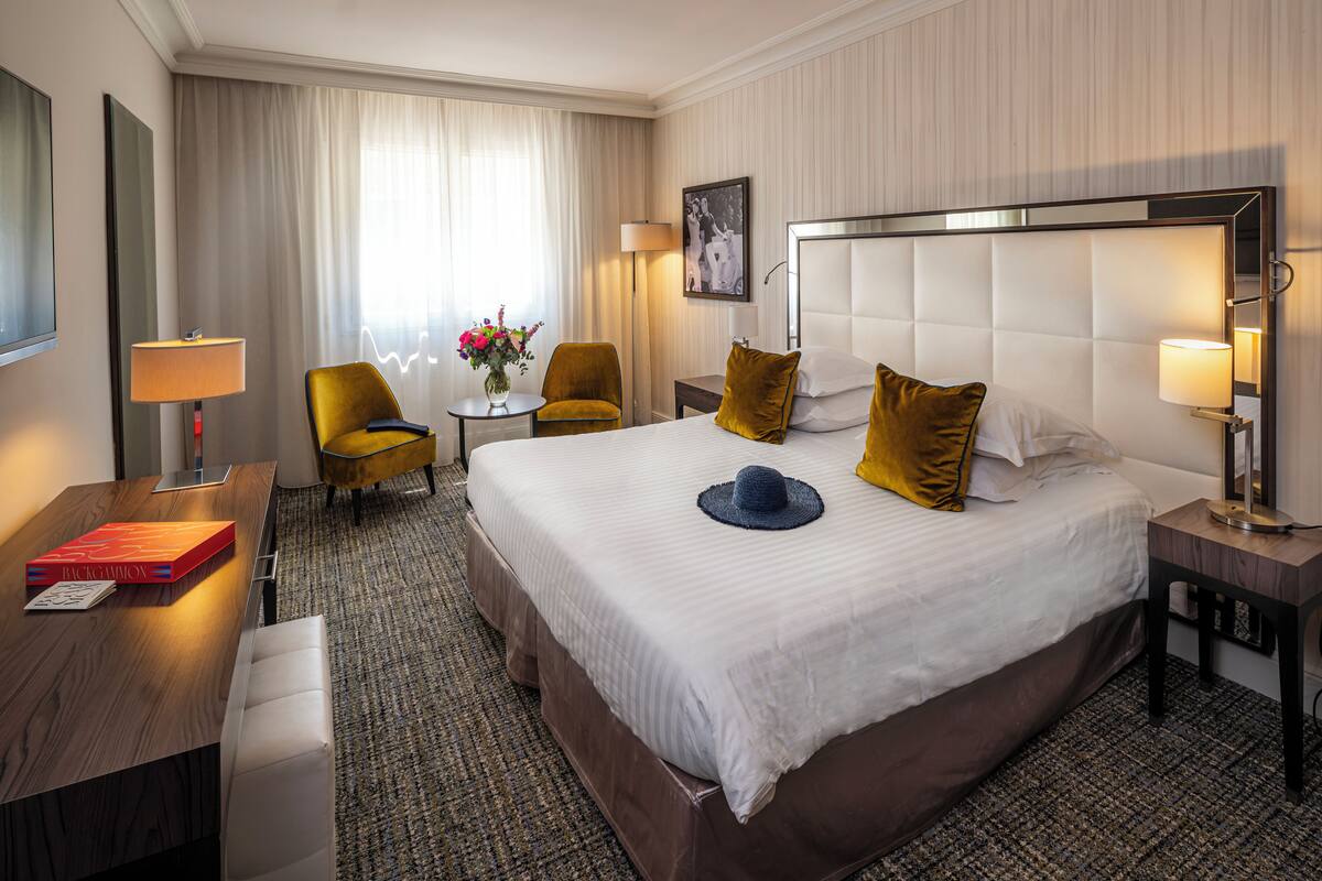 Een hotelkamer met een groot bed, een bureau met een laptop, een stoel, een kleintje tafeltje met een bloevas en een lamp.