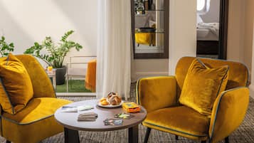 Une chambre d’hôtel avec deux fauteuils de couleur moutarde, une petite table ronde avec des magazines et un plateau de nourriture, une plante en pot et une vue sur un balcon avec un rideau blanc.