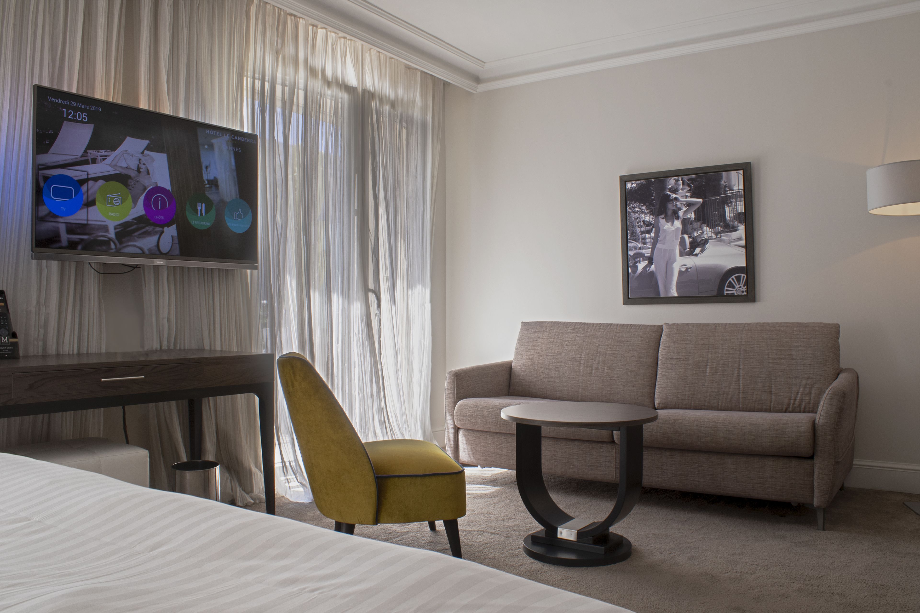 Una habitación de hotel con cama, escritorio, un sillón amarillo, un sofá gris, una mesita redonda pequeña, un televisor de pantalla plana y un cuadro en la pared.