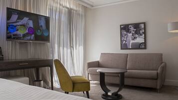 Una habitación de hotel con cama, escritorio, un sillón amarillo, un sofá gris, una mesita redonda pequeña, un televisor de pantalla plana y un cuadro en la pared.