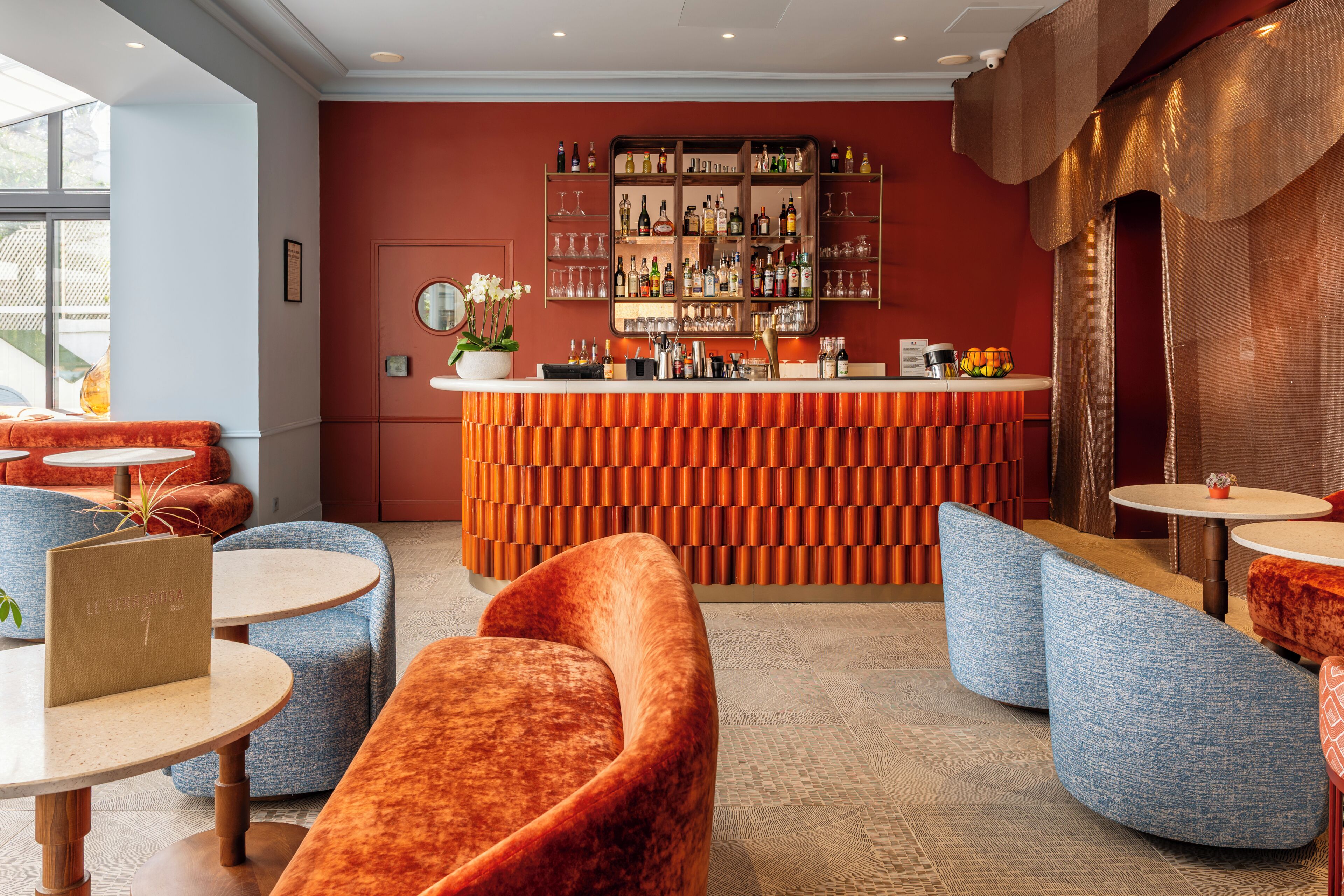 A bar area with a checkered orange and brown counter, blue upholstered seating, and a variety of bottles and glasses on shelves.