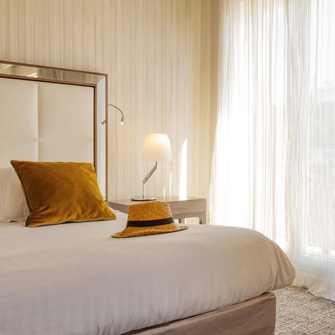 A hotel room with a bed, a bedside lamp, a mirror, and sheer curtains.