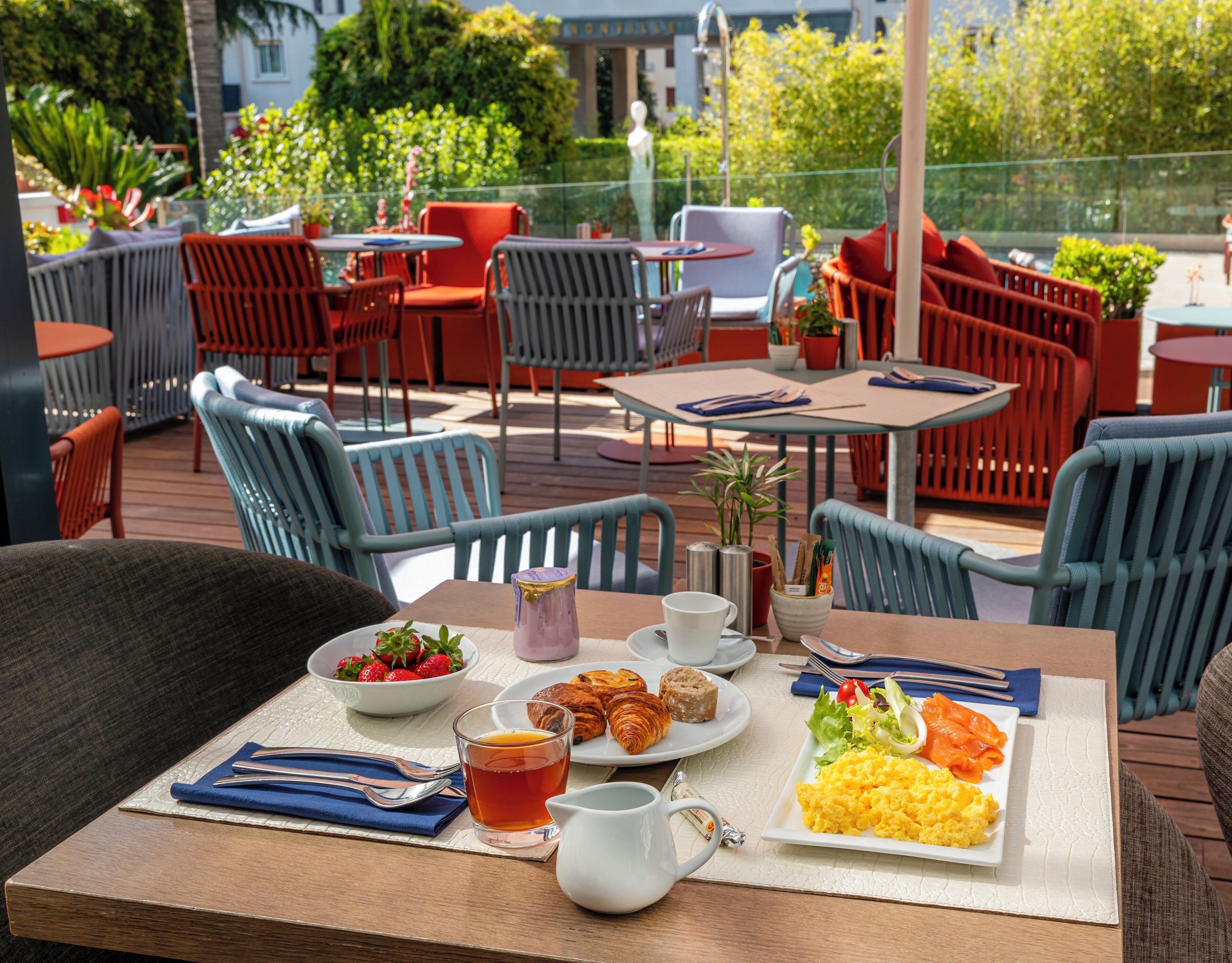 Ein Essbereich im Freien mit Tischen und Stühlen, ein Frühstücksbuffet mit Eiern, Toast und Obst sowie Blick auf Grünflächen und Gebäude im Hintergrund.