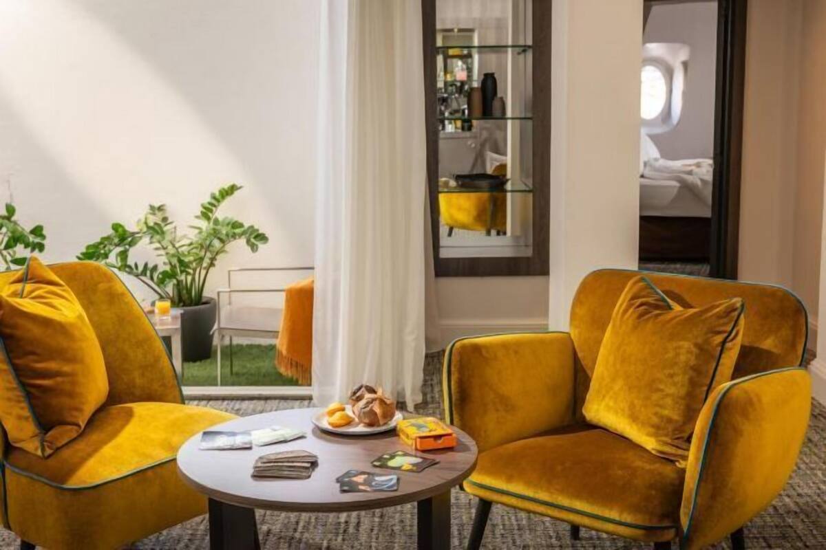 Une chambre d’hôtel avec deux fauteuils jaunes, une petite table ronde sur laquelle se trouve une assiette de nourriture, une bibliothèque et une fenêtre avec des rideaux.