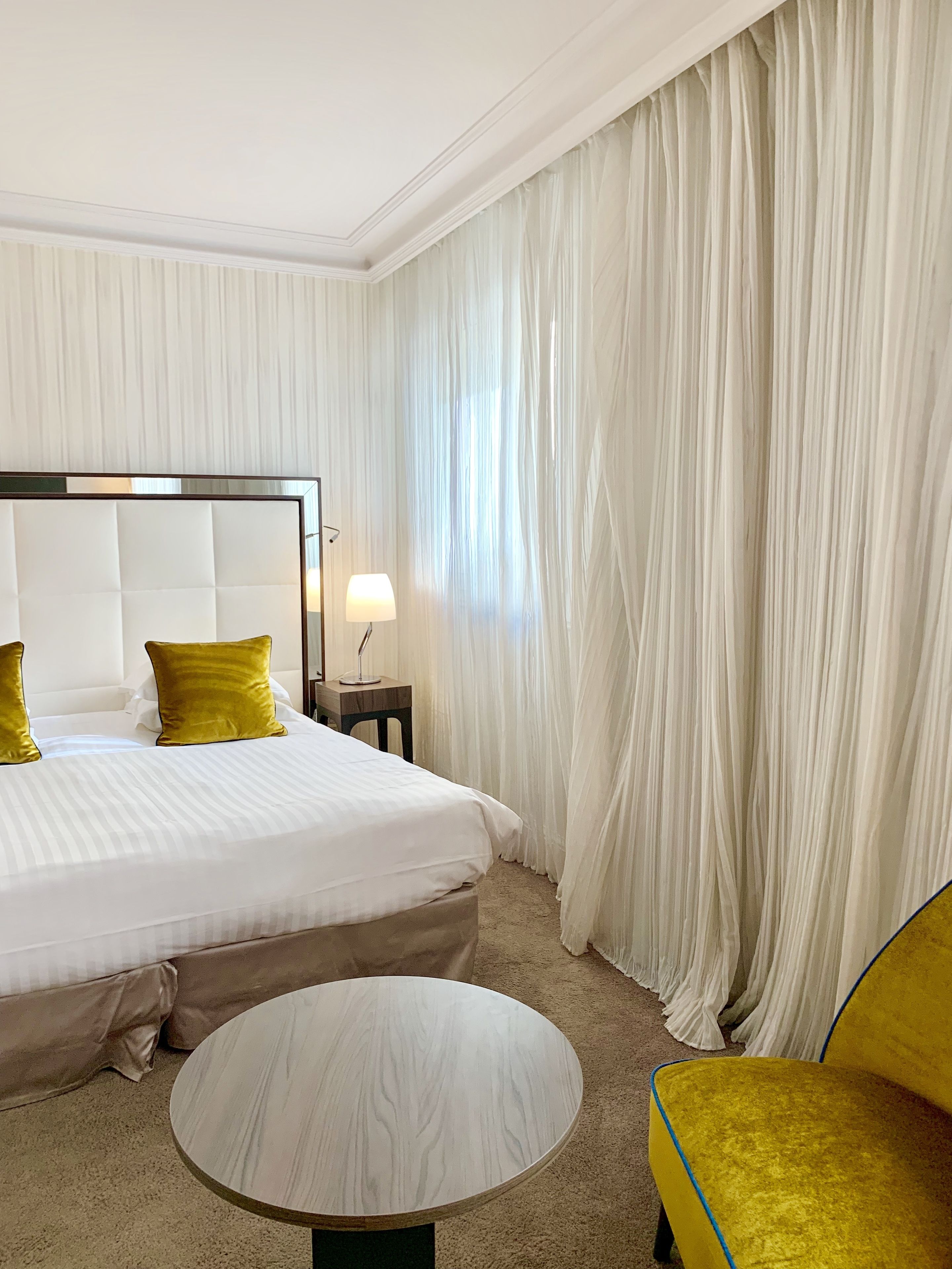 A hotel room with a large bed, a round table, a yellow chair, and a nightstand with a lamp.