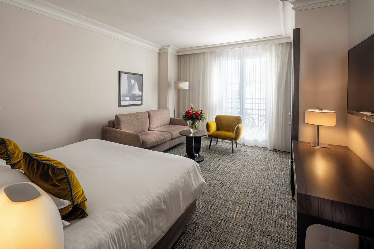 Une chambre d’hôtel avec un grand lit, un canapé, une petite table ornée de fleurs, un fauteuil jaune, un bureau avec une lampe et une télévision.