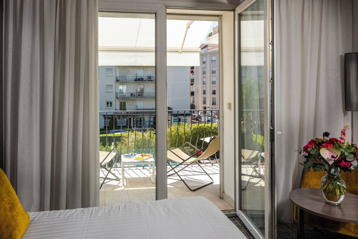 Une chambre d’hôtel avec un lit, une petite table ornée d’un vase de fleurs, un balcon avec des chaises et une table, et une vue sur des bâtiments et de la verdure.