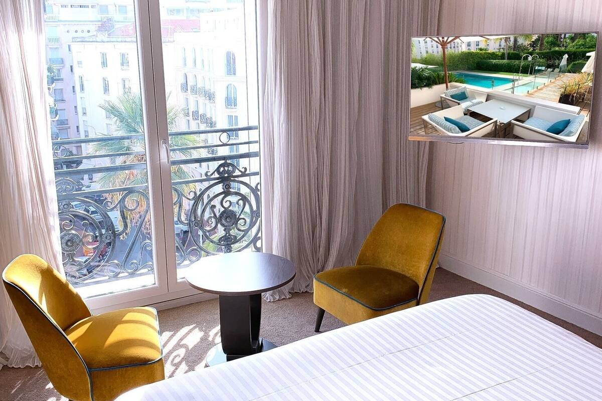 Une chambre d’hôtel avec un lit, deux fauteuils jaunes, une petite table ronde et une vue sur une piscine par la fenêtre.
