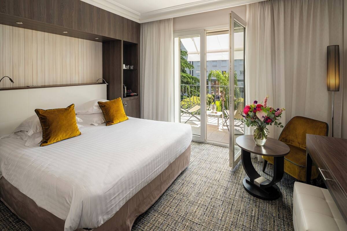 Une chambre d’hôtel avec un grand lit, une petite table sur laquelle est posé un vase de fleurs, une chaise et une vue sur l’extérieur.