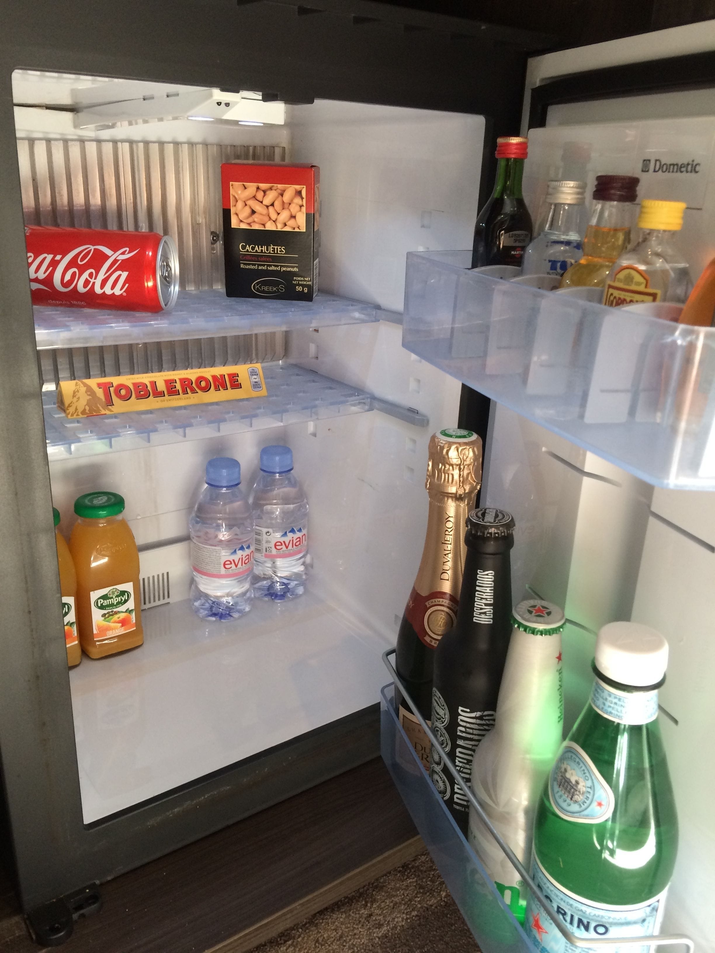 A refrigerator stocked with various beverages and snacks, including a can of Coca-Cola, Toblerone, Evian water, and Dom Pérignon champagne.