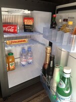 Un réfrigérateur rempli de diverses boissons et de collations, notamment une canette de Coca-Cola, une tablette de chocolat Toblerone, de l’eau Evian et une bouteille de champagne Dom Pérignon.
