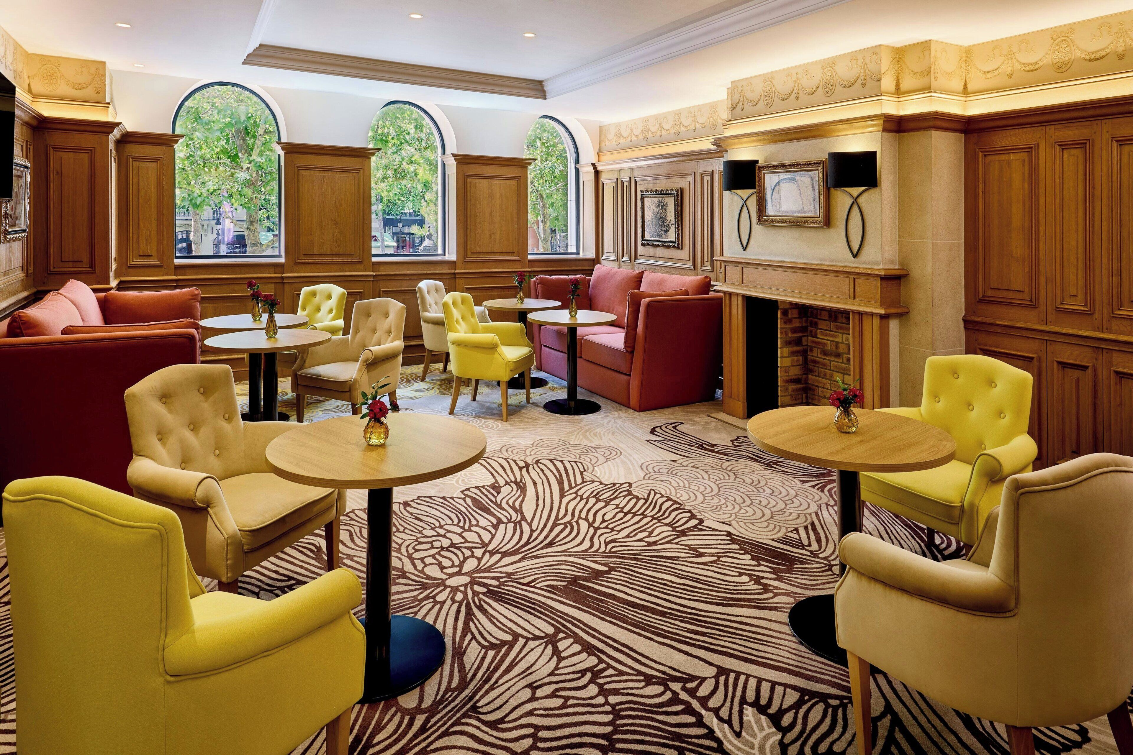 Un hall d’hôtel doté de plusieurs espaces salon, comprenant des canapés et des fauteuils jaunes et rouges, disposés autour de petites tables rondes. De grandes fenêtres en arc offrent une vue sur les arbres à l’extérieur.
