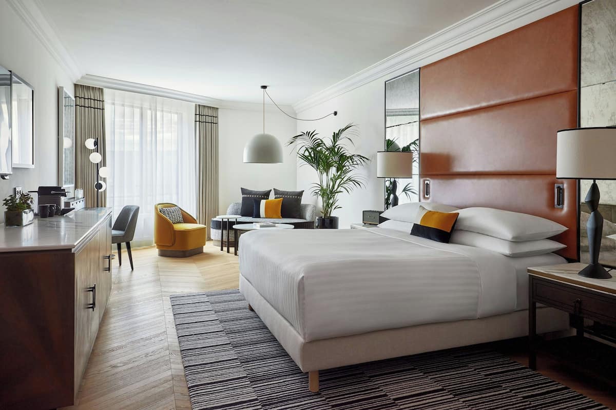 A modern hotel room with a large bed, a desk, a chair, a small table, and a potted plant.