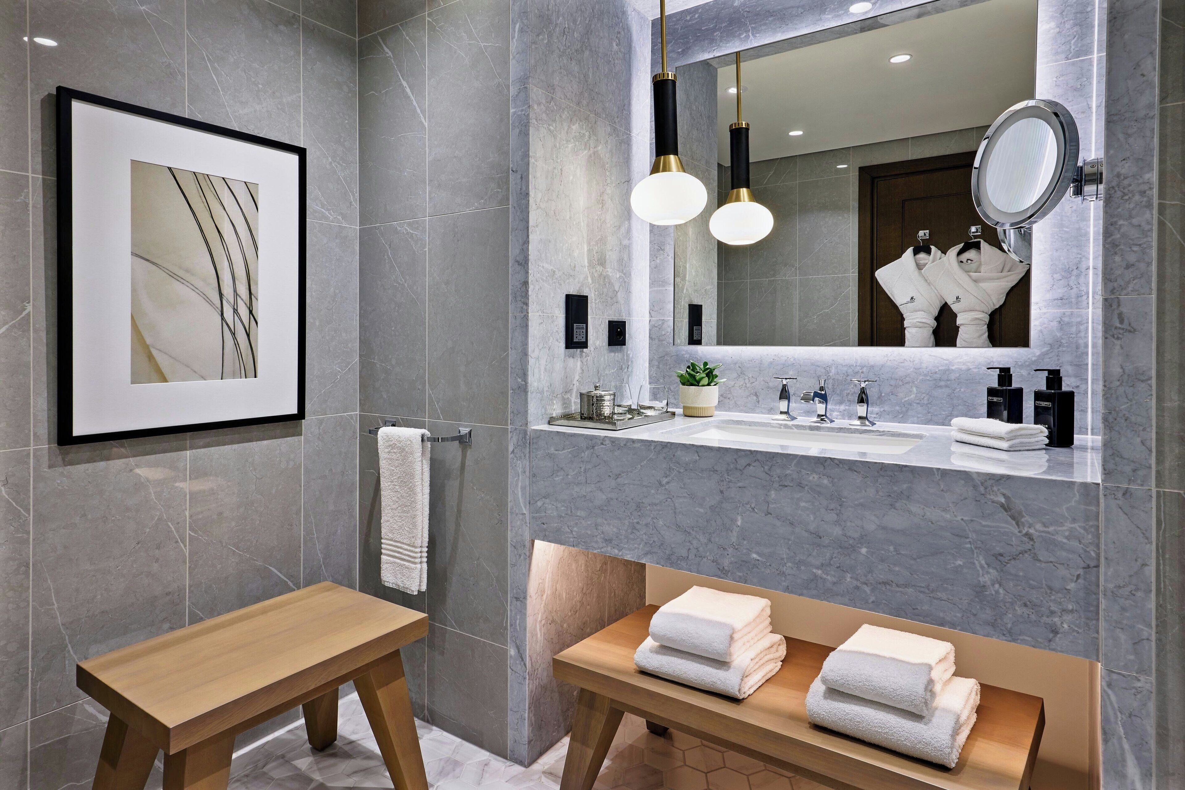 Un bagno moderno con parete in marmo, un'opera d'arte incorniciata, una panca in legno, un lavabo con specchio e asciugamani riposti con cura.