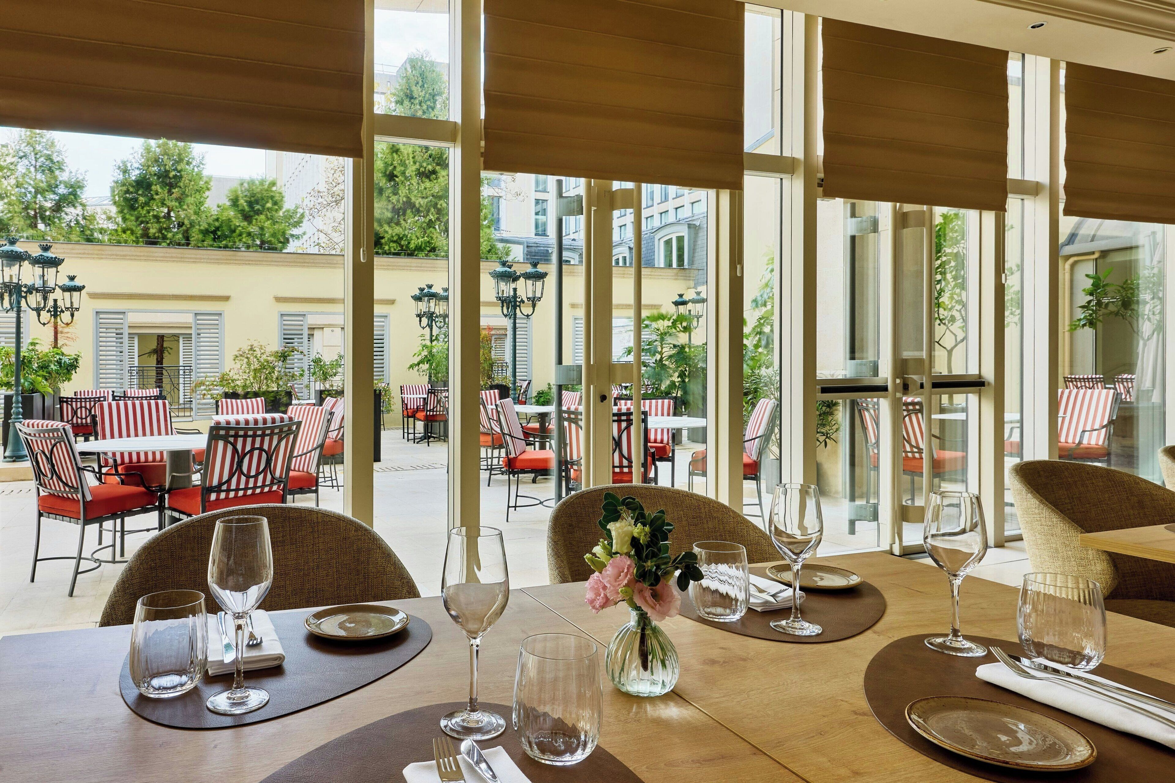 Un área de comedor al aire libre con mesas preparadas para dos, con vasos de vidrio transparente y un jarrón con flores.