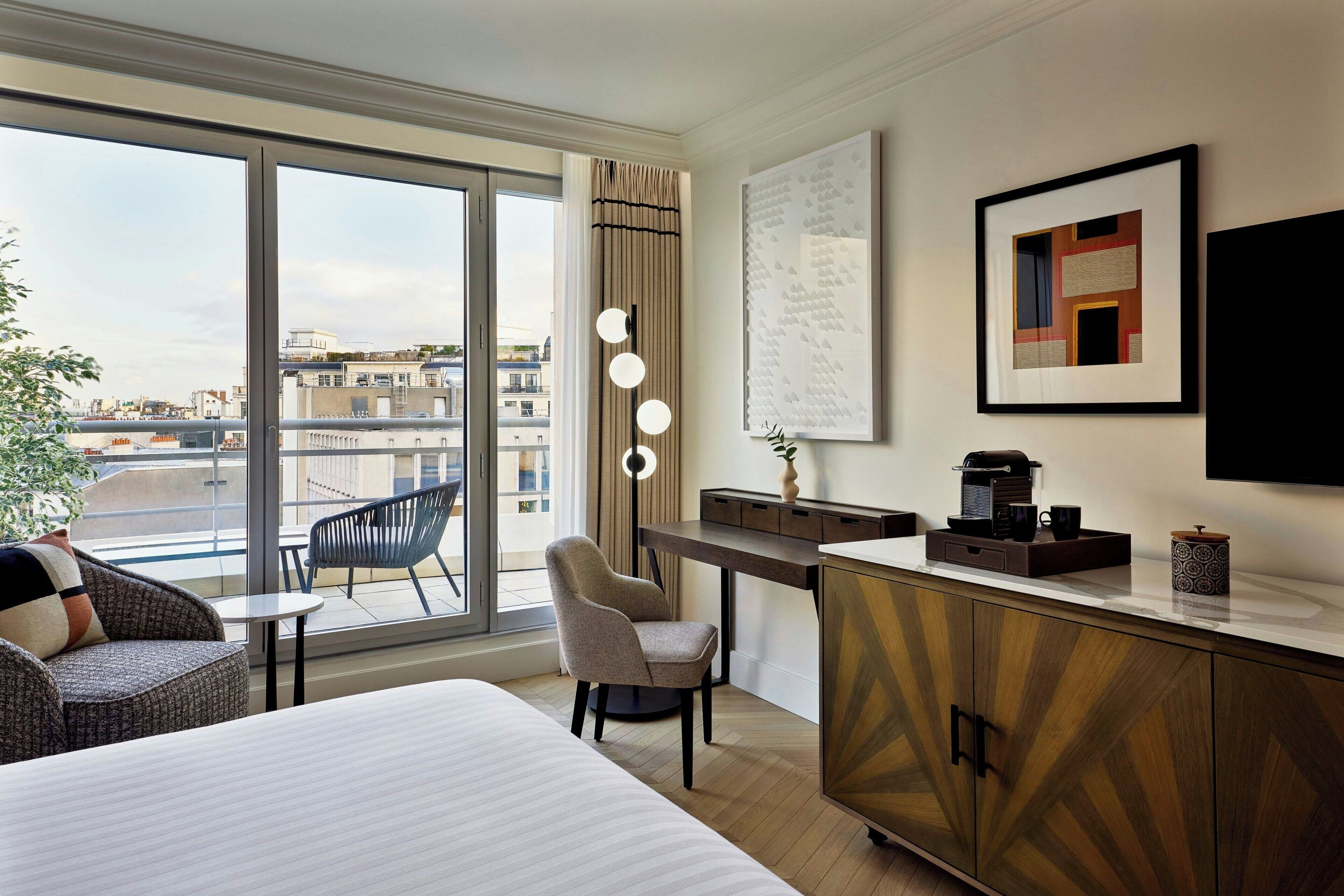 A hotel room with a large bed, a desk with a coffee maker, a chair, a small table, and a view of the city through large windows.