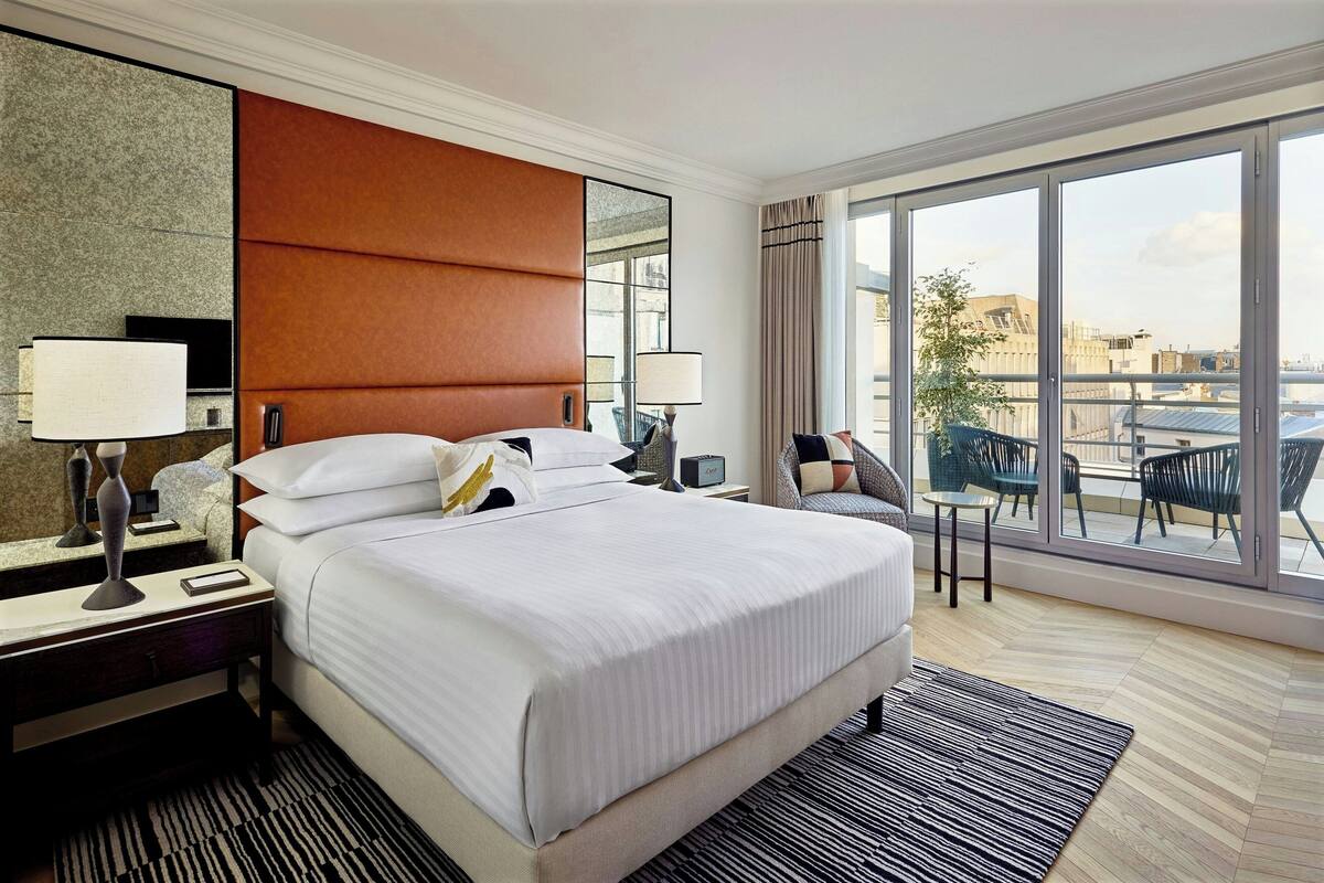 A hotel room with a large bed, a nightstand with a lamp, a small table, and a chair. There is a balcony with a view of the city.