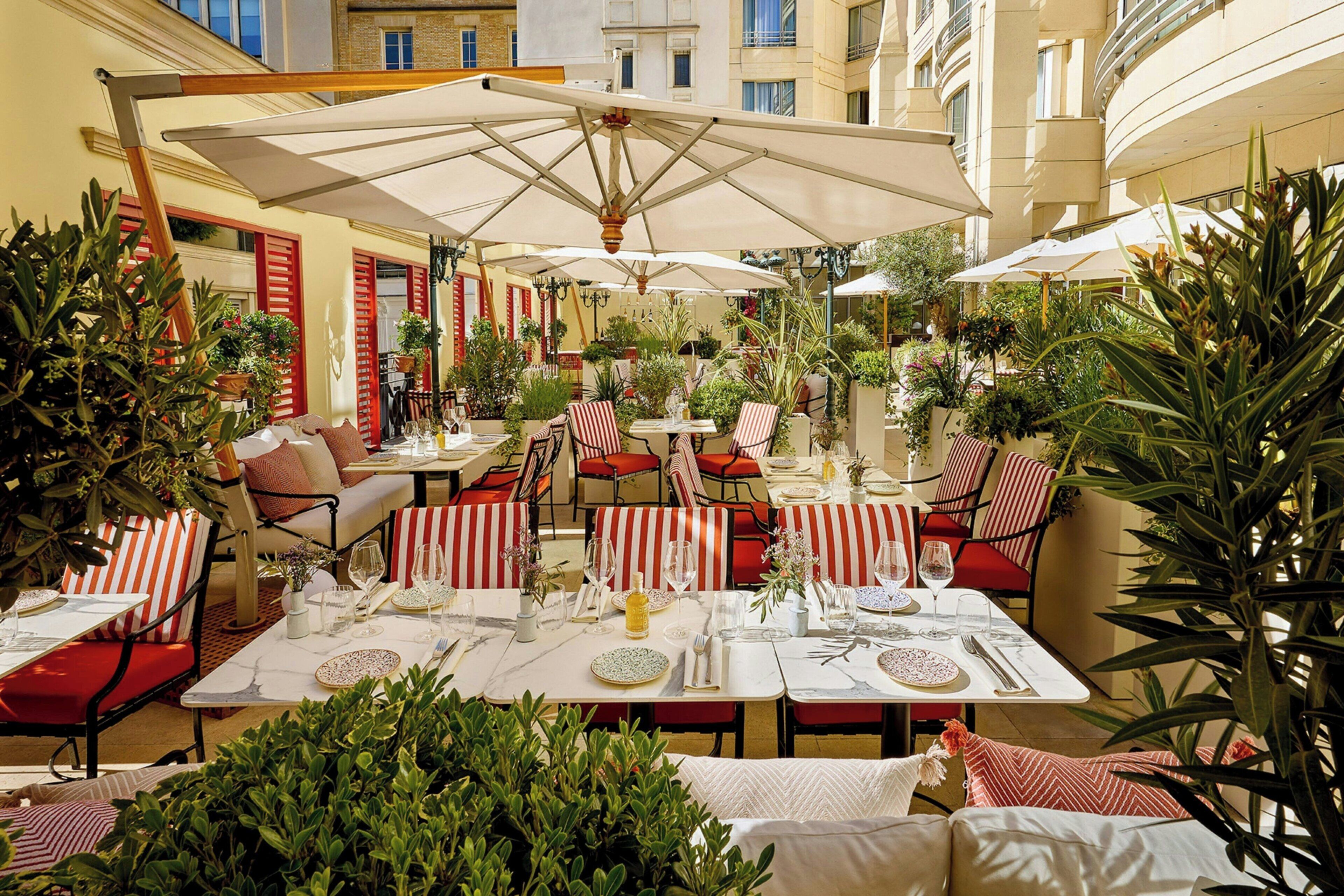 An outdoor dining area with tables, chairs, and umbrellas, surrounded by plants and red-striped cushions.