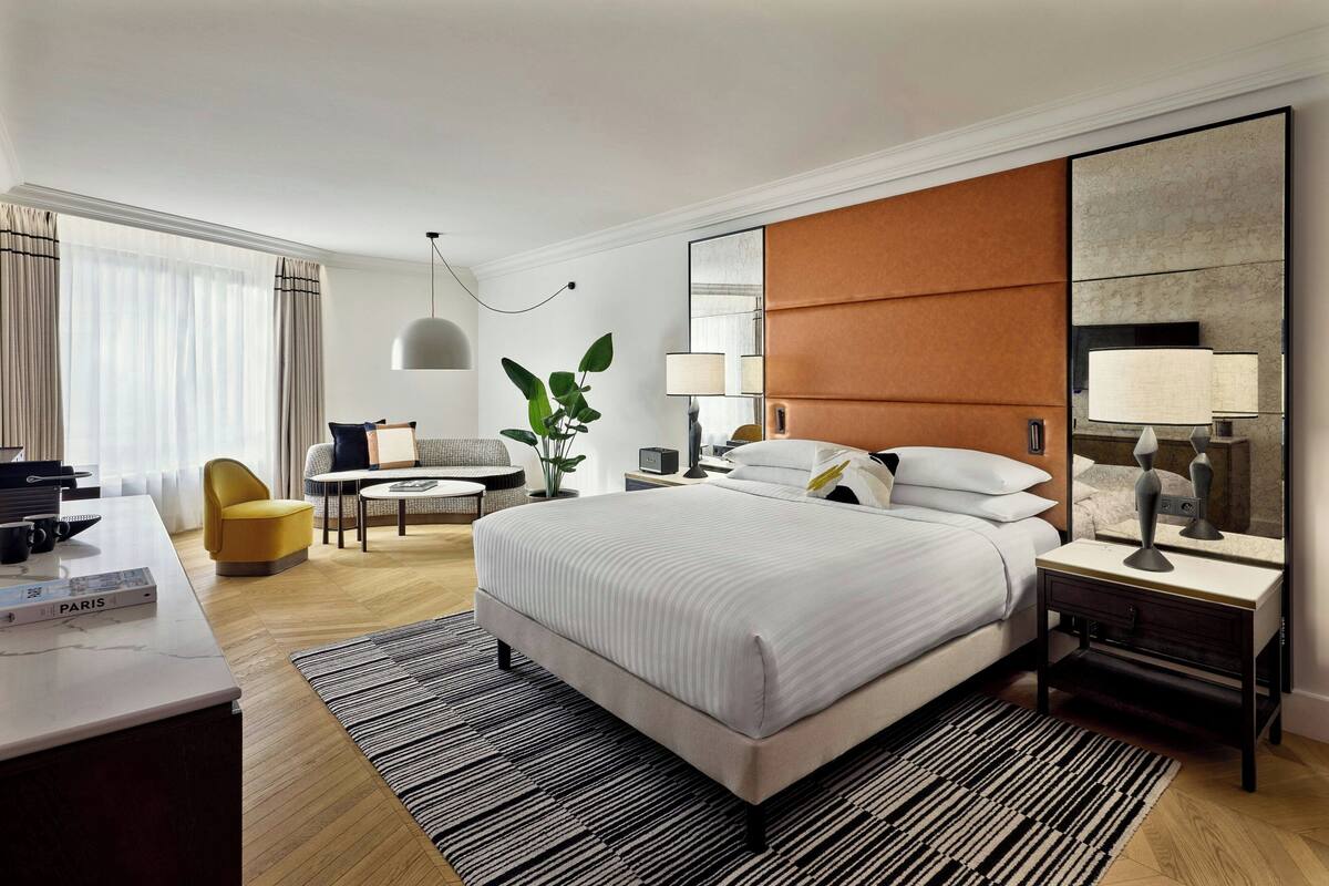 A modern hotel room with a large bed, a desk, a chair, a small table, and a plant.